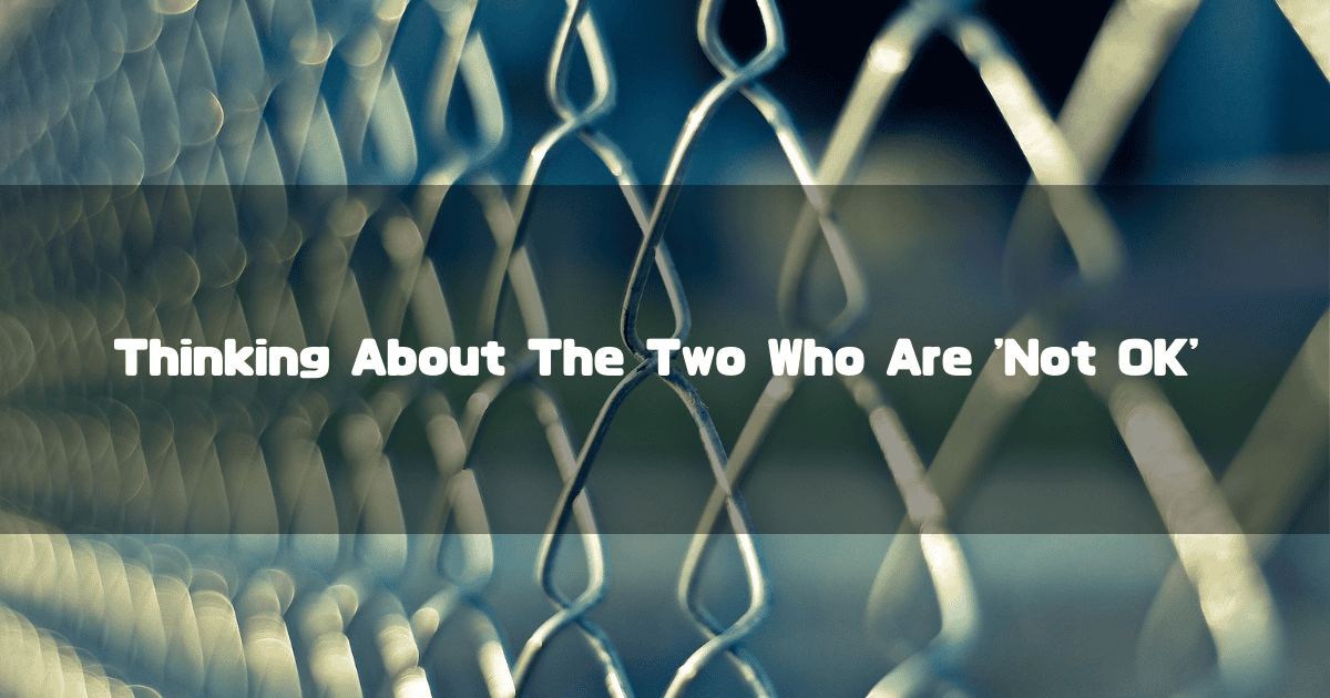 Close-up of a wire chain-link fence with a blurred background. Text reads: 'Thinking About The Two Who Are Not OK'.