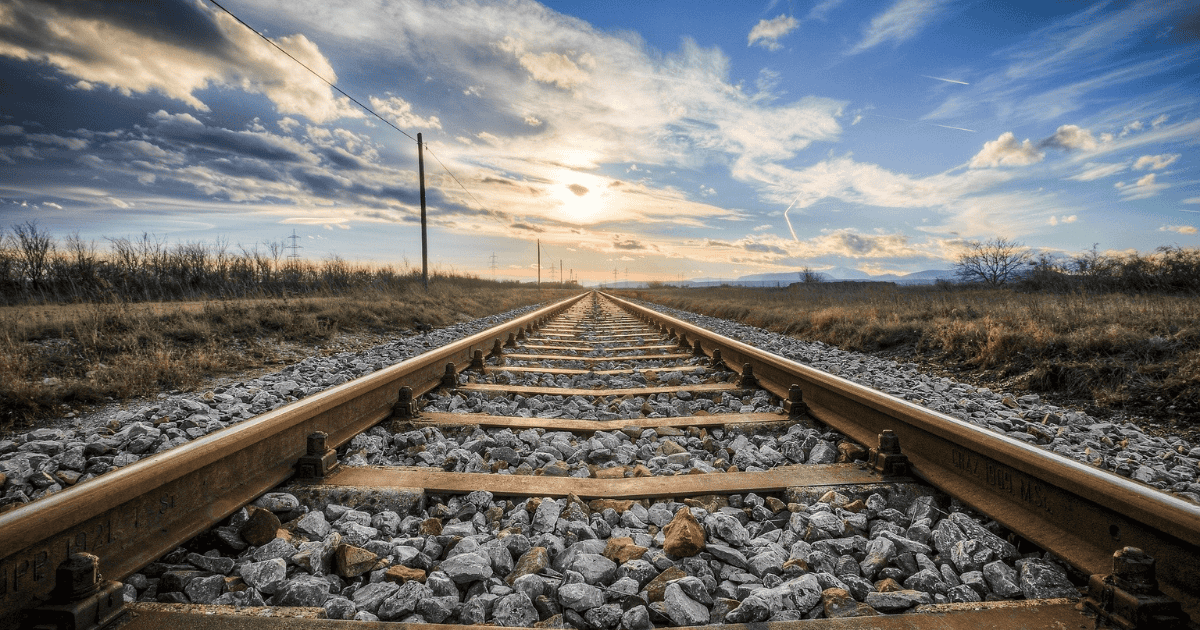 Railway tracks stretching toward the horizon through open countryside beneath a dramatic sunset sky